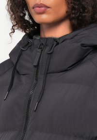 Casaco puffer preto com colarinho alto, apresentando um fecho de correr completo e fechos de cordão. Textura suave e detalhes de design minimalistas.