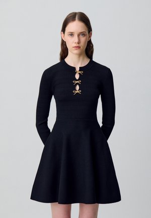 Donna con lunghi capelli castani, che indossa un vestito nero a coste con tre chiusure a forma di fiocco dorate sul petto, in piedi davanti a uno sfondo grigio chiaro uniforme.