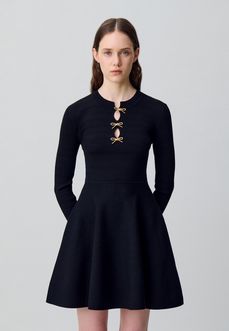 Donna con lunghi capelli castani, che indossa un vestito nero a coste con tre chiusure a forma di fiocco dorate sul petto, in piedi davanti a uno sfondo grigio chiaro uniforme.