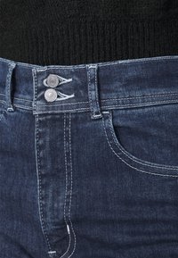 Jeans de denim azul oscuro con un diseño clásico de cinco bolsillos, dos cierres de botón plateados y costuras blancas en contraste.