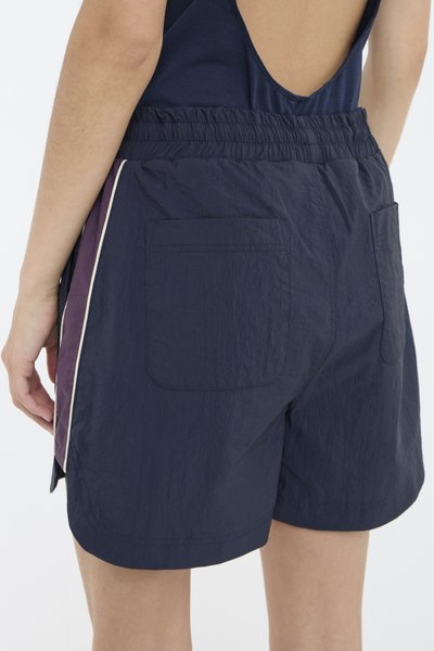 Marineblå shorts laget av lett materiale med elastisk linning, to baklommer og kontrasterende sidestykker i lilla.