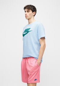 Νέος άνδρας με ανοιχτό μπλε t-shirt Nike και ροζ σορτς Nike, στέκεται με τα χέρια στις τσέπες μπροστά σε απλό φόντο.