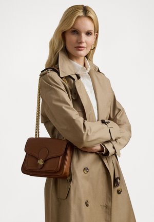 Brown leather shoulder bag with a flap closure, gold hardware, and embossed logo, paired with a beige trench coat and white shirt.