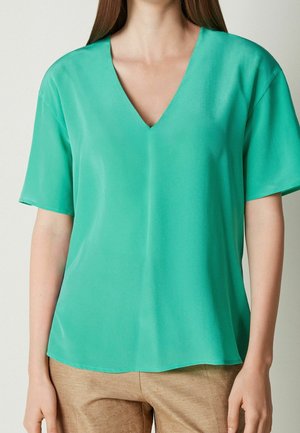 Kvinde iført en løs, kortærmet turkisbluse med V-hals og lysebrune bukser, stående foran en ensfarvet baggrund.