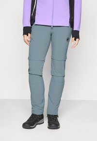 Persona che indossa pantaloni da trekking grigi con cerniera, scarponi da trekking neri e una giacca viola con maniche nere, in piedi su uno sfondo semplice.