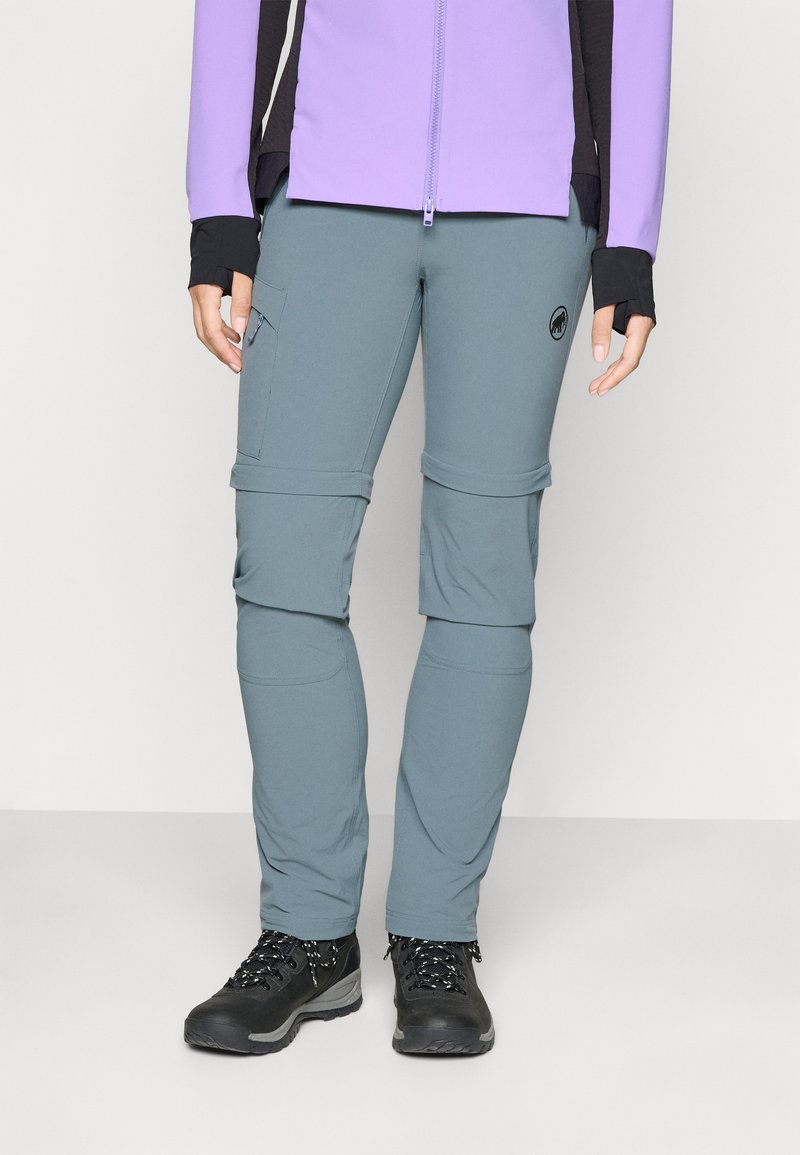 Persona che indossa pantaloni da trekking grigi con cerniera, scarponi da trekking neri e una giacca viola con maniche nere, in piedi su uno sfondo semplice.