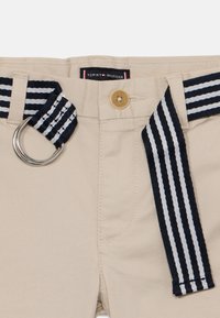 Pantalon en coton beige avec ceinture rayée marine et blanche, dotée d'une boucle argentée. Comprend une fermeture par bouton et une étiquette logo à la taille.