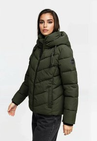 Kobieta w ciemnozielonej kurtce puchowej z wysokim kołnierzem i kapturem, stojąca na jednolitym jasnym tle.