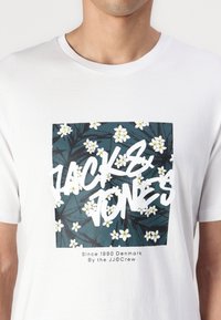 Bílý bavlněný tričko s čtvercovým grafickým motivem se zelenými listy a bílými květy, a textem "JACK & JONES" v tučném bílém písmu.