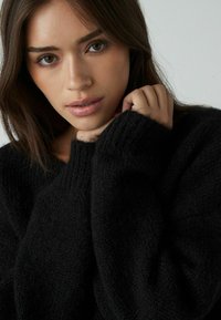Zwarte trui van zacht, textuurstof met een losse pasvorm, voorzien van oversized mouwen en een brede halslijn. Model steunt met de handen op de stof.