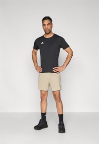 Zwart sportshirt met korte mouwen en wit Adidas-logo, gecombineerd met beige shorts en zwarte sportschoenen, wat een sportieve uitstraling geeft.