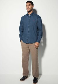 Donkerblauwe denim overhemd met lange mouwen en een knoopsluiting kraag, gecombineerd met beige broek en zwarte schoenen, tegen een lichte achtergrond.
