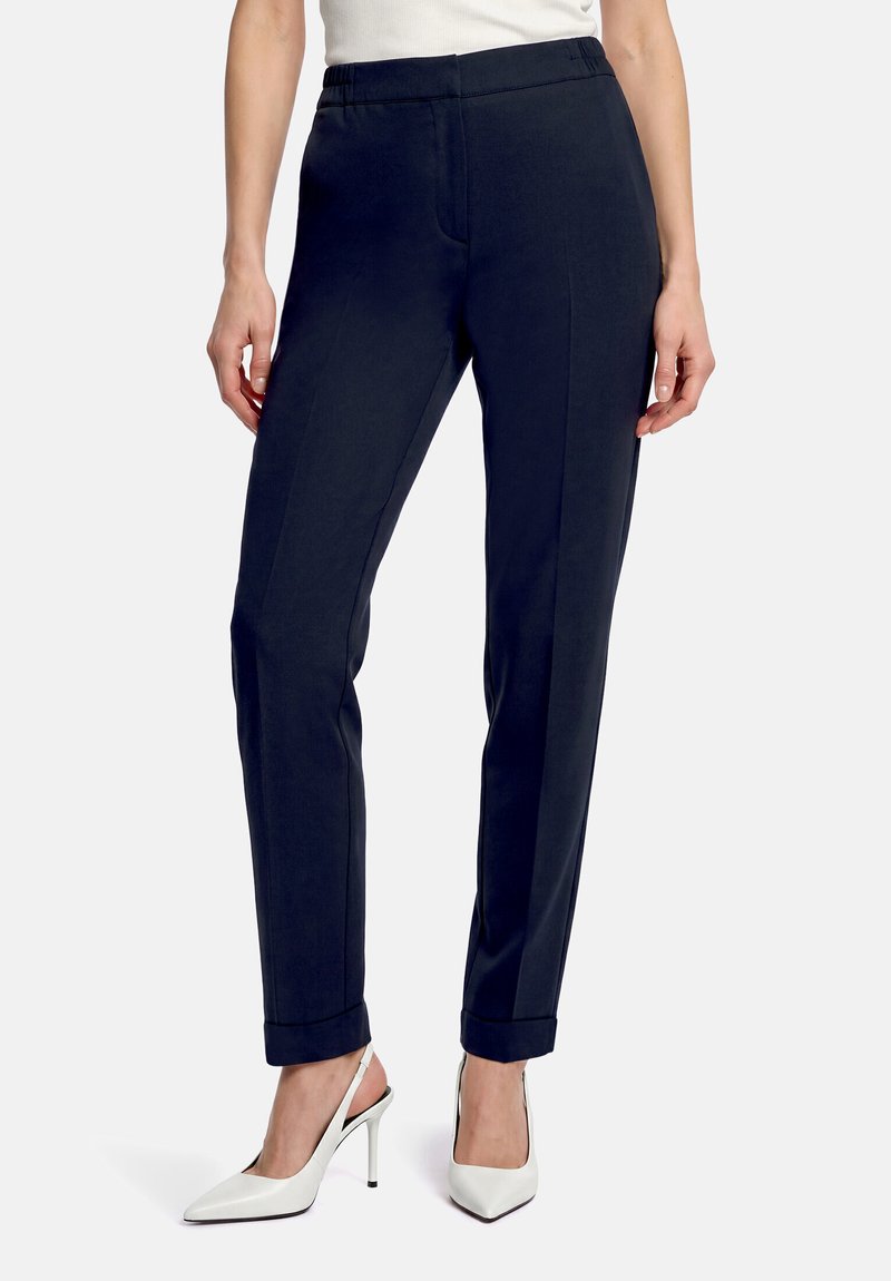 Femme portant un pantalon slim bleu marine avec ourlets retroussés et des chaussures slingback blanches à talons hauts pointus, debout sur un fond blanc.