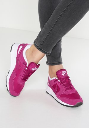 Personne portant des baskets Nike Air Pegasus rose vif et un jean noir, debout sur un sol blanc avec les jambes croisées.