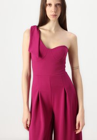 Fuksia jumpsuit med en-shoulder design, plisserte vide bukser og et tettsittende liv med subtile utskjæringer i utringningen. Jevn tekstur gjennom hele antrekket.