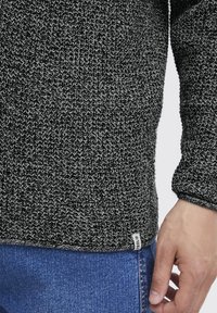Jersey de punto en blanco y negro con un patrón texturizado, que presenta una etiqueta en el dobladillo y un ajuste ceñido en los puños. Combinado con jeans de mezclilla azul.