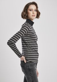 Sort og grå stribet langærmet skjorte med stående krave; blød tekstur og tætsiddende design. Båret med mørkejeans, hænderne på hofterne.