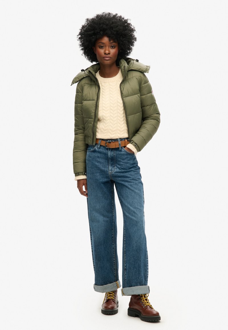 Olijfgroen pufferjack met capuchon, crèmekleurige kabelgebreide trui, losse blauwe jeans en bruine veterschoenen met gele accenten.