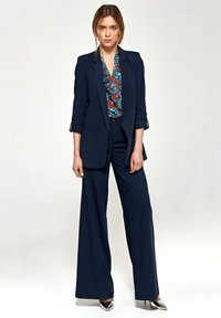 Granatowy garnitur z dopasowaną marynarką z podwiniętymi rękawami, w połączeniu z szerokimi spodniami i wzorzystą bluzką w niebiesko-czerwonych odcieniach.