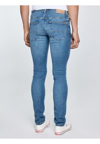 Žmogus, dėvintis siaurus mėlynos spalvos džinsus ir baltus sportbačius, stovintis priešais vientisą šviesią foną, žiūrint iš nugaros.