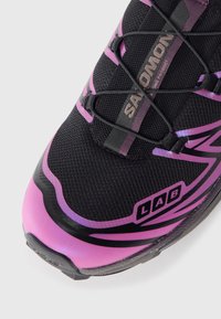 Svarte atletiske sko med mesh-struktur, med rosa aksenter, logo og elastiske lisser. Bøyd såle med en svak overgang fra svart til rosa.
