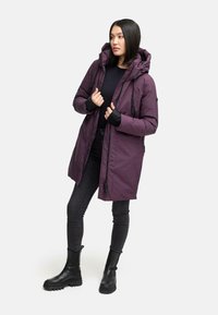 Lilla polstret parka-jakke med hætte, lynlås foran, sidelommer og sorte ærmekanter, parret med sorte skinny jeans og støvler.