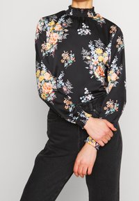 Blouse noire à fleurs avec des manches longues, un col froncé et des motifs floraux multicolores, dotée de poignets élastiques. Texture de tissu lisse.