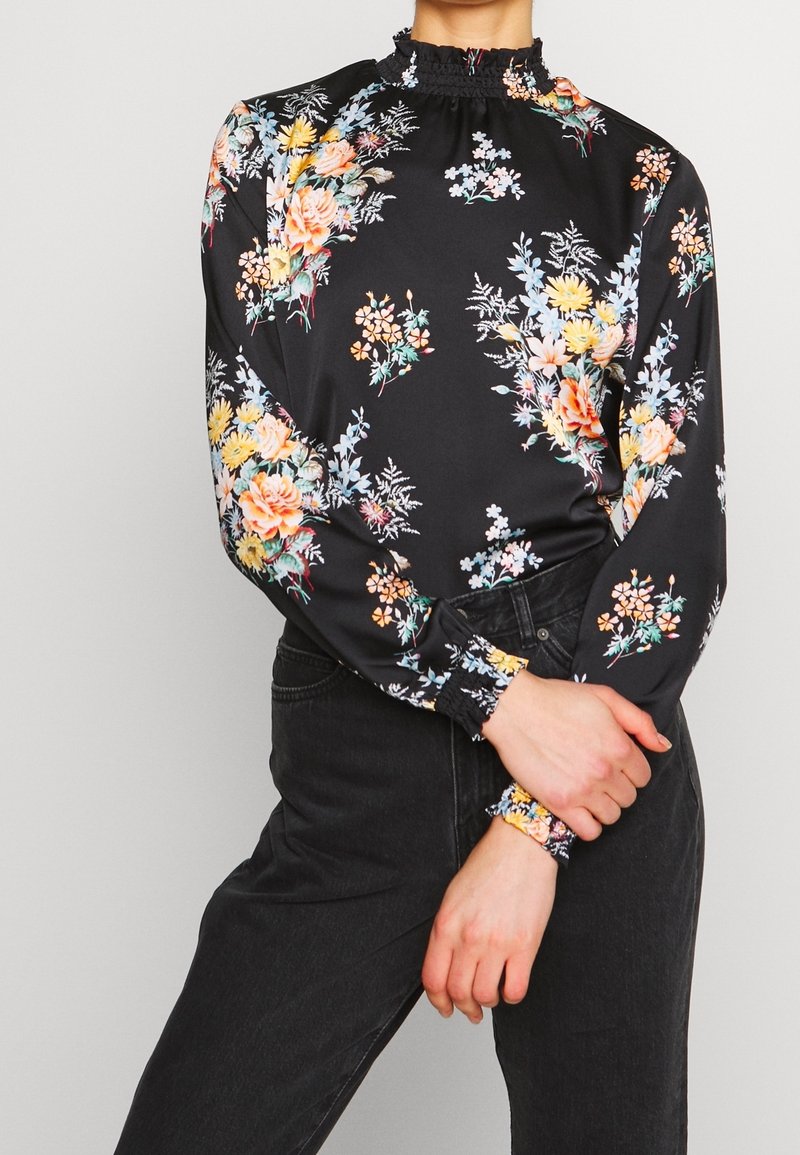 Blouse noire à fleurs avec des manches longues, un col froncé et des motifs floraux multicolores, dotée de poignets élastiques. Texture de tissu lisse.