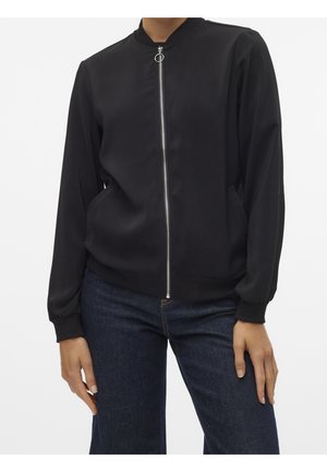 Kvinde iført en sort bomberjakke med ringlynlås og mørkeblå jeans, stående mod en ensfarvet hvid baggrund.