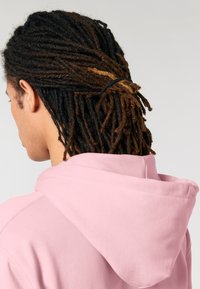 Felpa rosa con un grande cappuccio, realizzata in un tessuto morbido e con cuciture visibili, indossata da un individuo con lunghe trecce acconciate all'indietro.