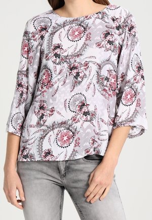 Femme portant un chemisier ample à manches longues à fleurs rose, gris et blanc, associé à un jean gris clair.