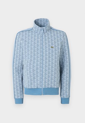 Ανοιχτό μπλε μπουφάν με φερμουάρ, γεωμετρικό μοτίβο, ριμπ μανσέτες και τελείωμα, και μικρό πράσινο λογότυπο κροκόδειλου στο στήθος.