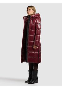 Vínově červená prošívaná bunda s kapucí, zapínáním na zip vpředu a postranními kapsami. Hladký, lesklý materiál s délkou sahající ke kolenům. Nošena přes černé oblečení.