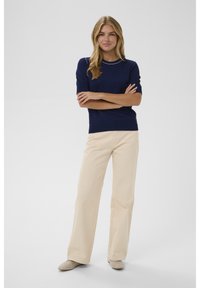 Moteris, stovinti su sulenktais rankomis, vilkinti tamsiai mėlyną trumpomis rankovėmis marškinėlį, kreminės spalvos plačiajuosčius kelnes ir kreminės spalvos batus su naginėmis ant baltos fono.