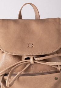 Tan leather backpack with a fold-over flap, drawstring closure, and front zipper pocket. Features subtle branding and contrast stitching.