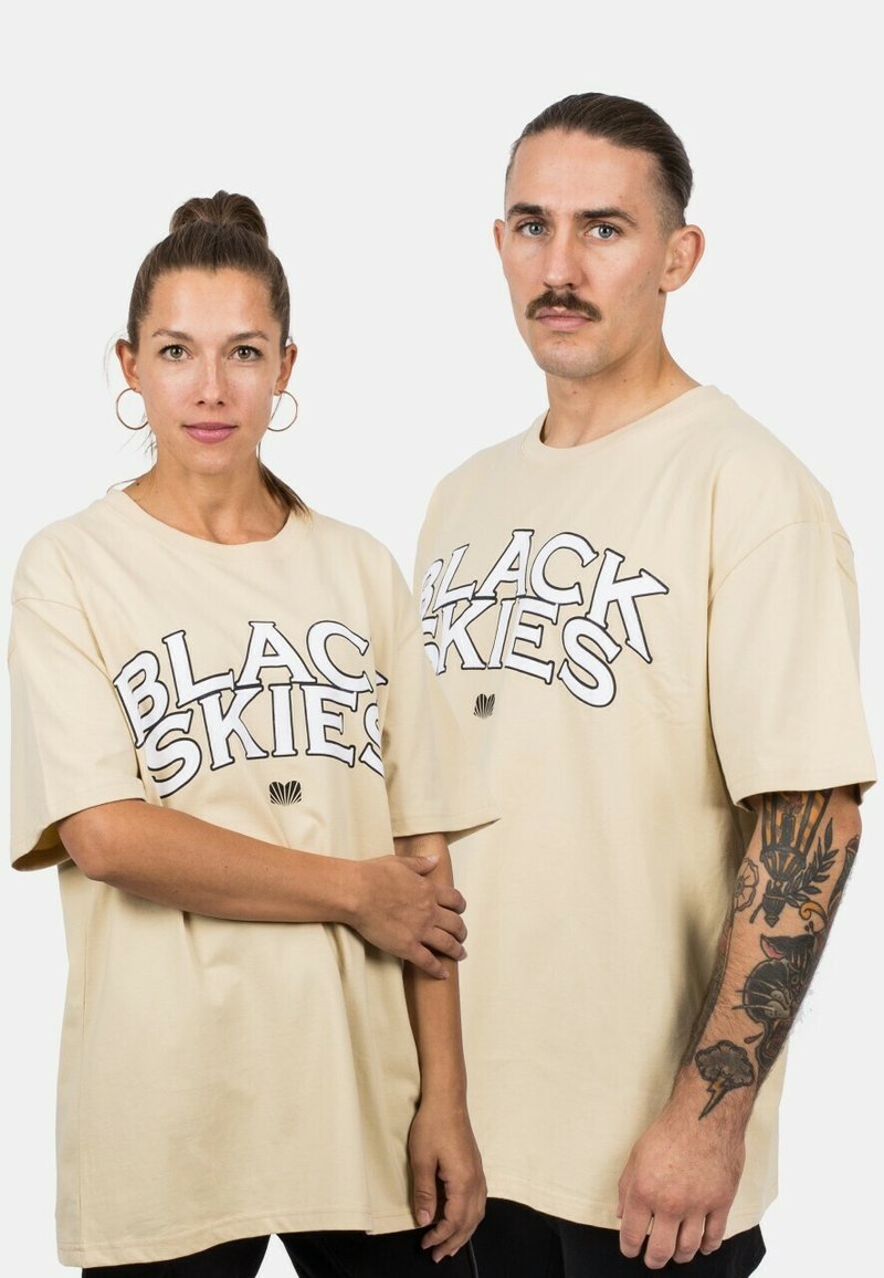 Beige, túlnyúló pólók, melyeken vastag fekete betűkkel van feltüntetve a "BLACK SKIES" felirat. Puha pamut anyag, rövid ujjak és laza fazon.