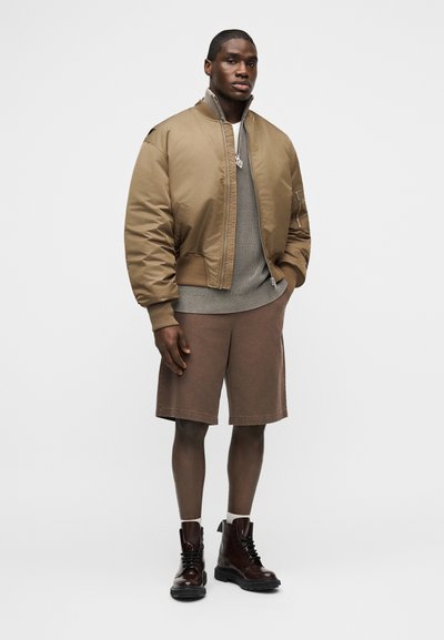 Άνδρας που φοράει μπεζ bomber μπουφάν, γκρι πλεκτή μπλούζα, καφέ σορτς, λευκές κάλτσες και σκούρα καφέ μποτάκια με κορδόνια, στέκεται μπροστά σε απλό φόντο.