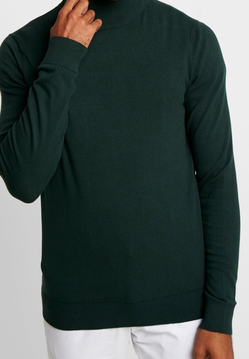 Pull à col roulé vert foncé en matériau doux et fin tricoté, avec col et poignets côtelés. Présente une coupe ajustée.