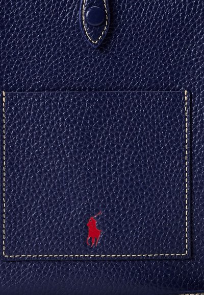 Primo piano di pelle testurizzata blu navy con una tasca cucita e un piccolo logo rosso di giocatore di polo al centro in basso.