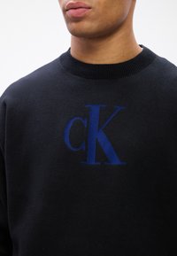 Čierna mikina s širokým rebrovaným výstrihom a veľkým modrým vyšitým logom "cK" na hrudi. Mäkka textúra, ležérny dizajn.