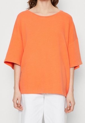 T-shirt surdimensionné orange vif à manches courtes, col rond, et texture en coton lisse. Associé à un pantalon blanc pour le contraste.