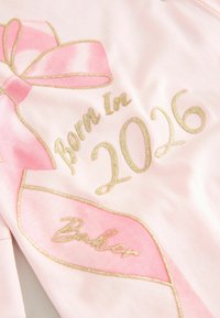 Heledad roosa kangas, millel on kuldse tikandiga tekst "Born In 2026" ja "Baker", koos roosa lipsu kujunduse ja sädelevate aktsentidega.