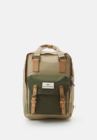 Backpack with tan body and green front pocket. Features leather straps, logo patch, and dual top handles. Sturdy fabric with visible stitching.