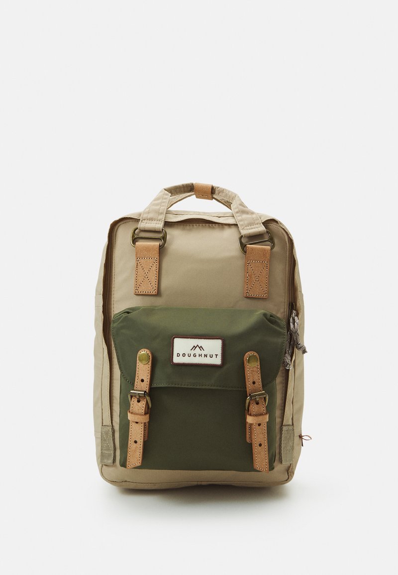 Backpack with tan body and green front pocket. Features leather straps, logo patch, and dual top handles. Sturdy fabric with visible stitching.