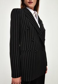 Nainen, joka käyttää mustaa viivallista kaksirivistä bleiseriä, jossa on kuusi nappia, valkoisen kauluspuseron päällä, näkyy vyötäröstä ylöspäin.