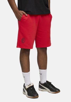 Shorts de sport rouges en tissu doux, dotés de poches latérales et d'un logo noir brodé. Portés avec des baskets noires et des chaussettes blanches.