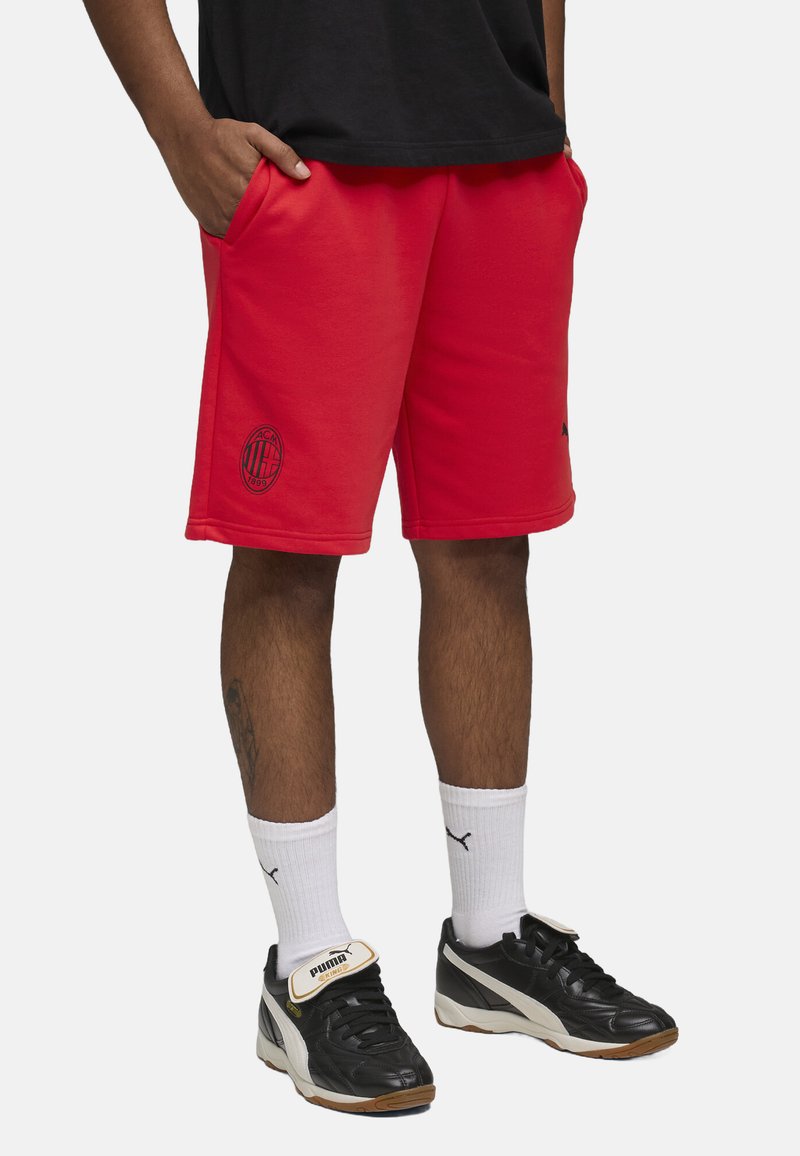 Pantalones cortos deportivos rojos hechos de una tela suave, con bolsillos laterales y un logo negro bordado. Combinados con zapatillas negras y calcetines blancos.