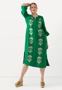 Grünes Leinenkleid mit goldener Blumenstickerei entlang der Mitte und der Ärmel, mit seitlichen Schlitzen und einem kragenlosen Ausschnitt. Getragen mit beigen Plateauschuhen.