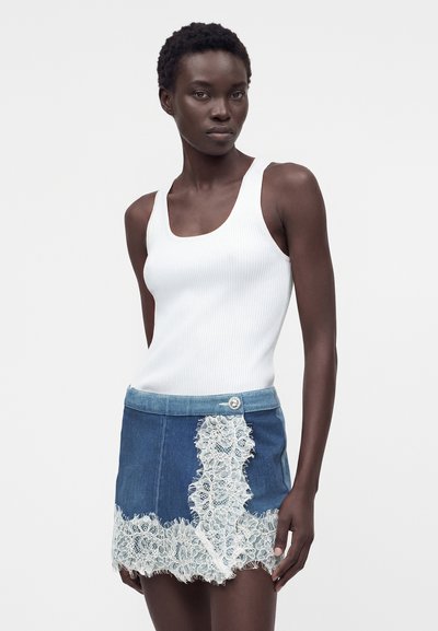 Γυναίκα που φοράει λευκό ριμπ τοπ και μπλε τζιν μίνι φούστα με λευκές δαντελένιες λεπτομέρειες σε απλό φόντο.