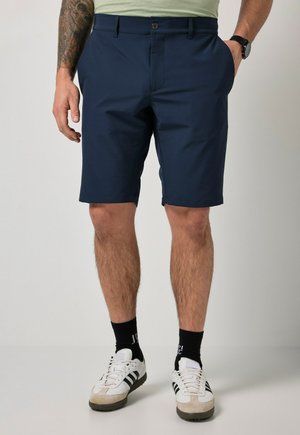 Mand iført marineblå shorts, sorte sokker og hvide sneakers med sorte striber, stående mod en ensfarvet baggrund.